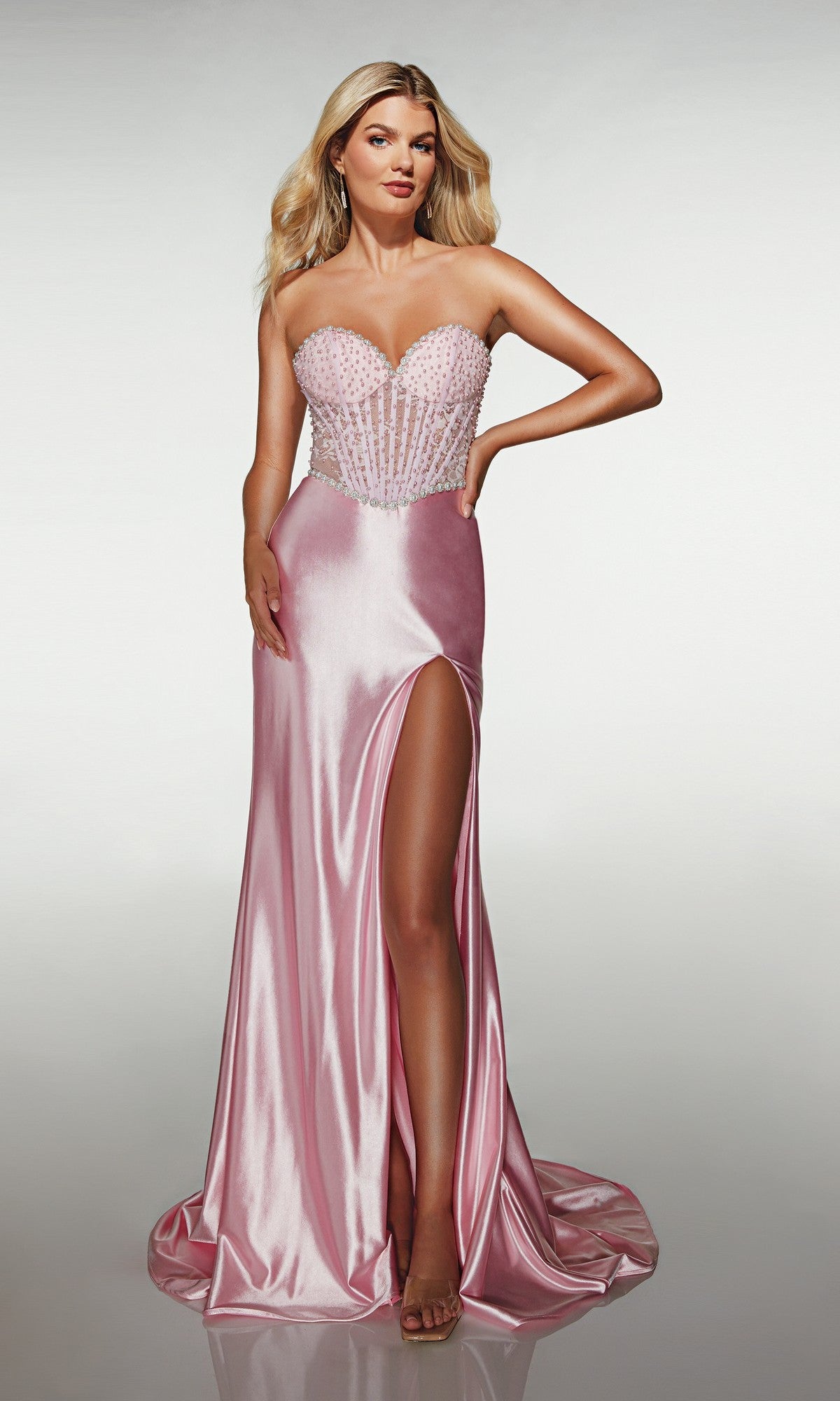 A woman models the Alyce 62209 long prom dress in light pink, featuring a strapless satin design, beaded corset bodice, and high-leg slit, as she poses against a plain light background.